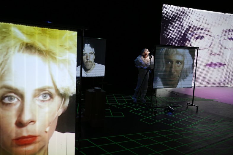 On several levels of the picture there are screens on which the faces of the performers of the stage can be seen. One person stands between the screens and their face is also projected onto one of the screens.
