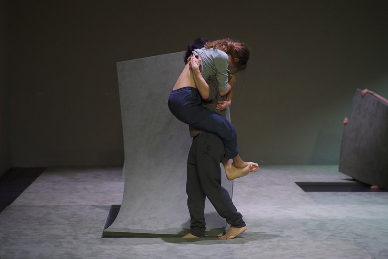 Photography of a stage scene. A person clings to another person. She is being carried by her. Large elastic floor panels are lifted from the floor.