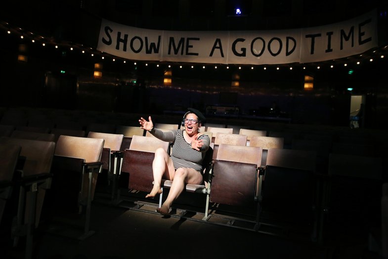 A woman sits in HAU1. Above her hangs a handwritten banner with the words "Show me a good time".