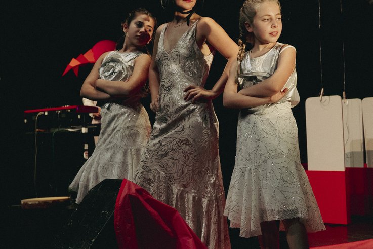 Drei Personen stehen in silbernen Kostümen auf der Bühne. Zwei jüngere Mädchen stehen mit verschränkten Armen neben einer Frau, die in der Mitte einen silbernen, glitzernden Helm trägt.