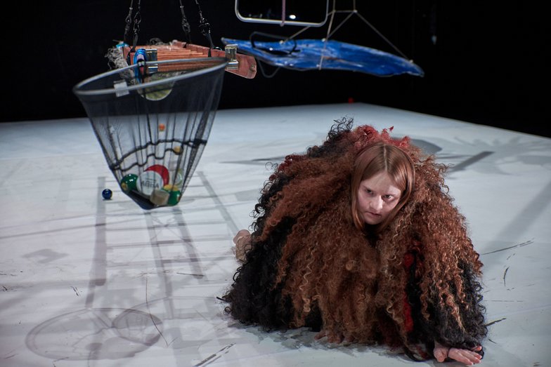 A person crawls on all fours on the floor. She wears a poncho made of long curly wool. A flight object, a ladder and a net filled with billiard balls hang from the ceiling around them.