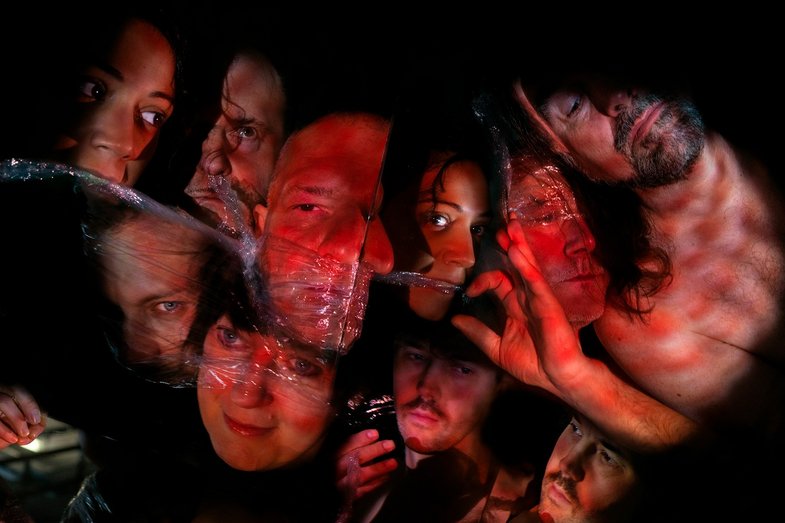 10 mit Frischhaltefolie bedeckte Gesichter sind in einem roten Licht auf ungewöhnliche Weise angeordnet. Auf dem Bild sind drei Hände zu sehen.