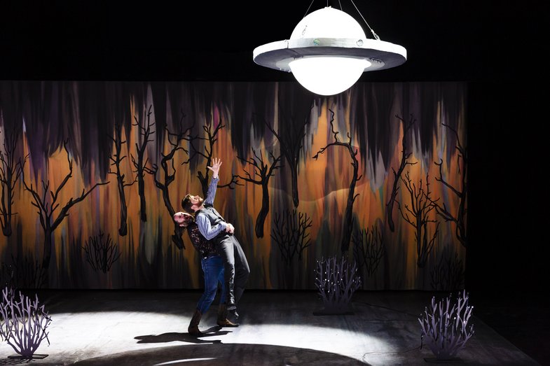 Zwei Personen auf der Bühne, eine hebt die andere von hinten hoch, der Arm ist nach oben in Richtung eines UFOs gestreckt. Im Hintergrund ist ein brennender Wald dargestellt.