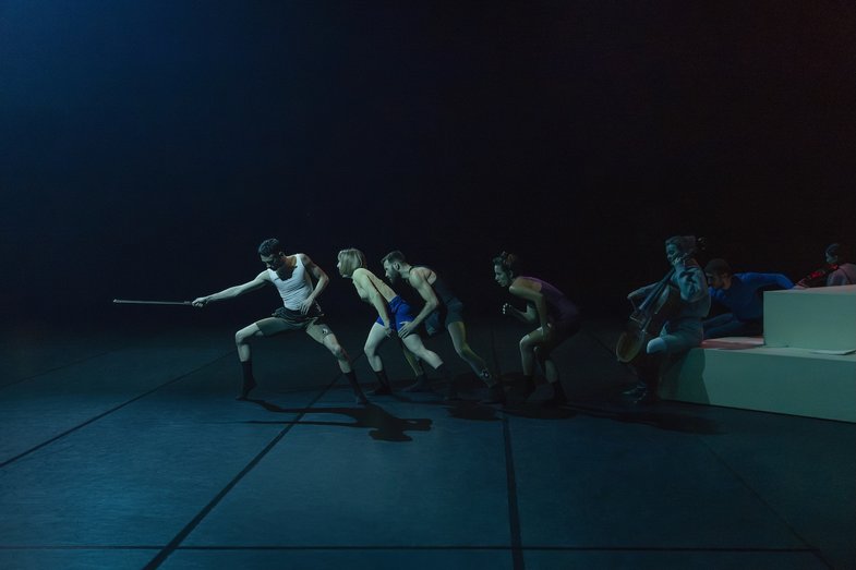 Vier Performer:innen in dunklem Licht ziehen gemeinsam an einem Stock, während Musiker:innen im Hintergrund auf einer stufenförmigen weißen Plattform sitzen.