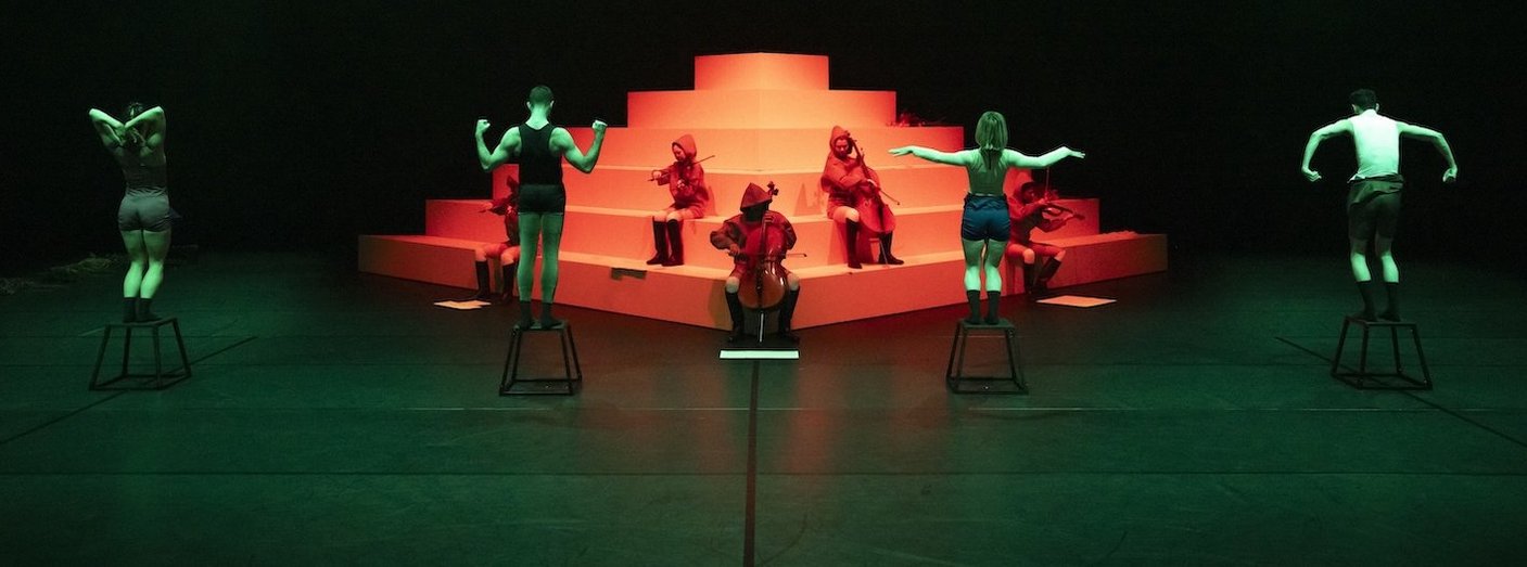 Performer:innen auf Hockern und Musiker:innen in roten Kapuzen posieren vor einer stufenförmigen weißen Plattform im grünen und roten Licht.