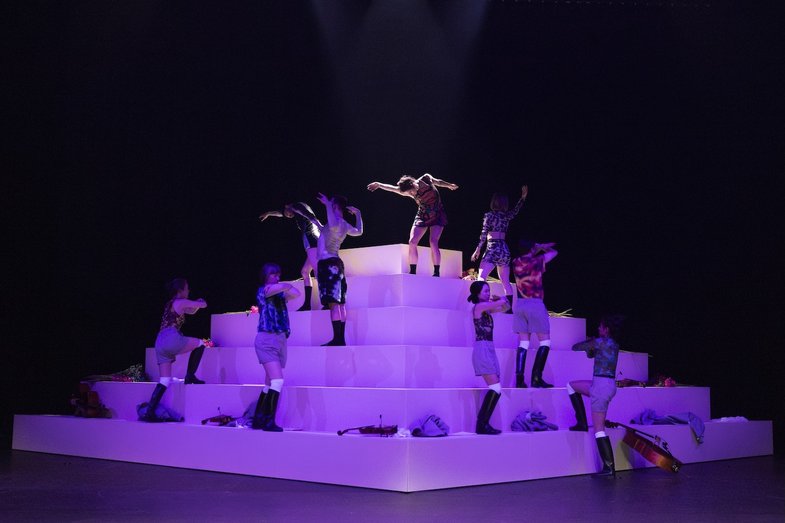 Performer:innen bewegen sich asynchron auf einer stufenförmigen weißen Plattform unter violettem Licht, umgeben von Kleidung und Instrumenten.