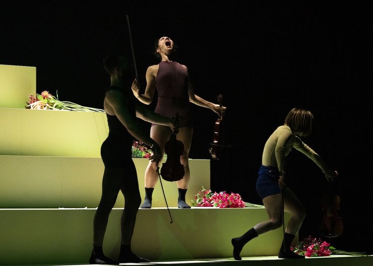 Drei Performer:innen mit Geigen auf einer stufenförmigen weißen Plattform unter grünem Licht, umgeben von Blumen, eine Person schreit expressiv mit offenem Mund in die Luft.