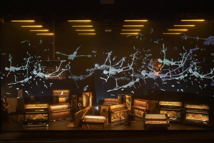 Photograph of a stage on which numerous upright pianos have an open case so that the hammer mechanism is free. Human skeletons hover above them.