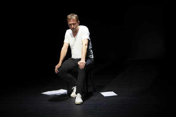 Boris Nikitin sits on the chair in an empty stage space in a thoughtful attitude. The Din A 4 pages of the text presented lie to his left and right.