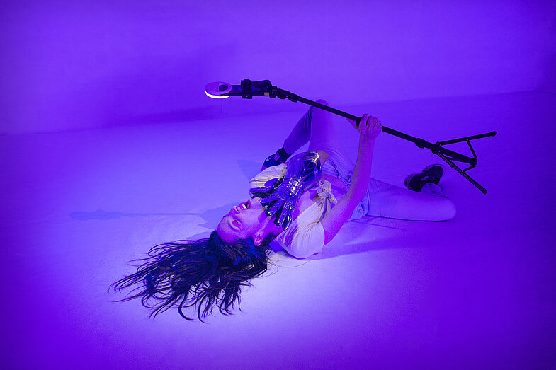 Photograph of the choreographer and dancer Helena de Laurens during the performance _jeanne_dark_. She is lying on the floor in a white room. In her right hand she holds a rig to which a smartphone with a ring light is attached. She stages herself for the camera. She has slipped a glove of armor over her left hand. She strokes her neck with it. The scene is lit with violet light.