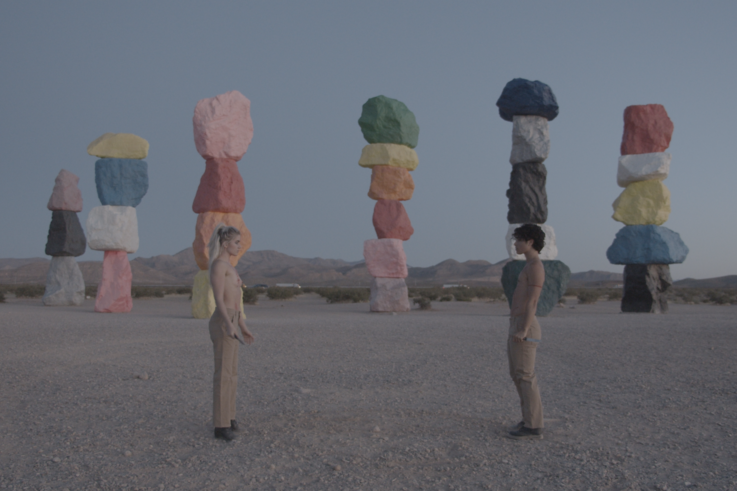 The two people face each other shirtless on a square in the mountains. In the background are colored boulders stacked on top of each other in a row.