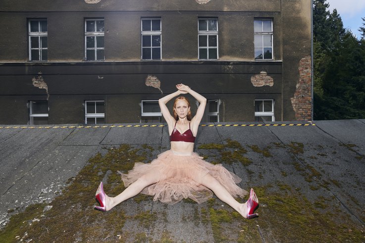 Photograph of Marianna Simnett on a flat roof. She is sitting in a tulle skirt in an open pose. The arms are stretched in the air. Looking towards the camera.