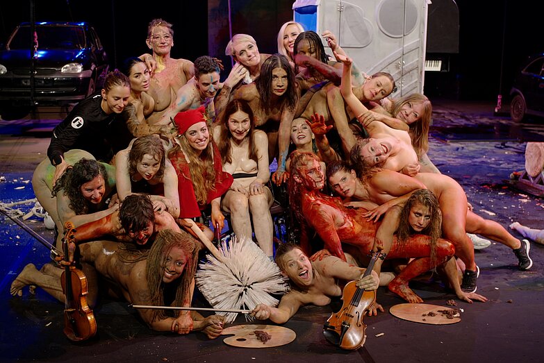Gruppenfoto auf der Bühne. Die Körper sind teilweise nackt und voller Farbe. Zwischen den Personen sind Requisiten wie eine übergroße Klobürste oder eine Violine.