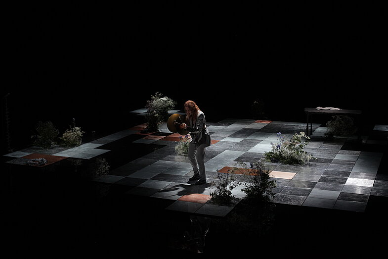 The dancer and choreographer Kat Válastur stands on the stage with the flowers and has a percussion instrument in her hand.