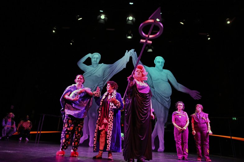 Five people are standing on the stage, the person in the middle is holding a kind of long stick towards the camera. In the background are two ancient Greek-looking large figures made of paper.