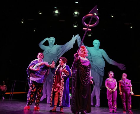 Five people are standing on the stage, the person in the middle is holding a kind of long stick towards the camera. In the background are two ancient Greek-looking large figures made of paper.
