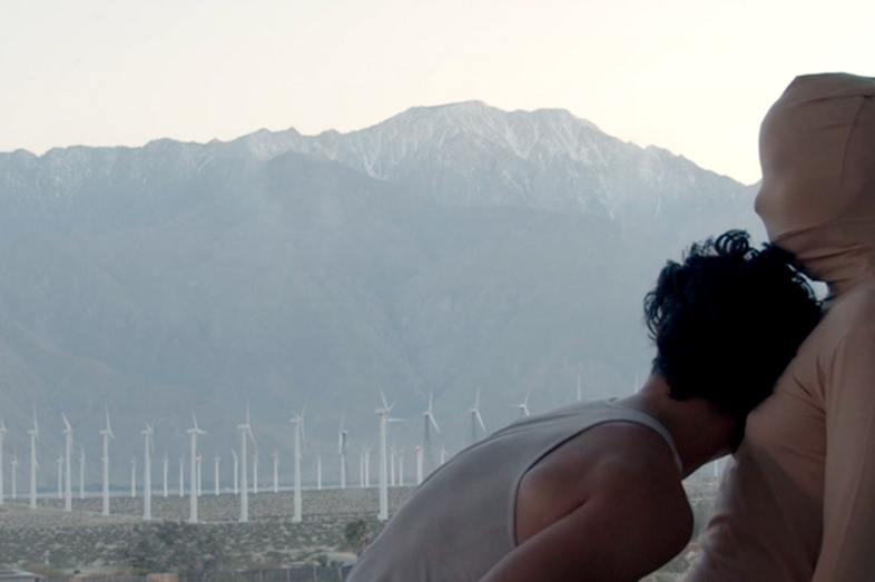 One person has their head on the other person's chest. In the background stretches a mountain landscape and a wind farm.