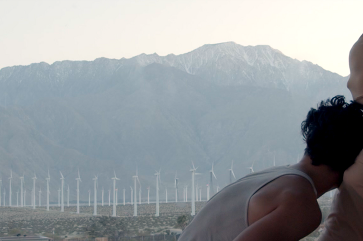 One person has their head on the other person's chest. In the background stretches a mountain landscape and a wind farm.