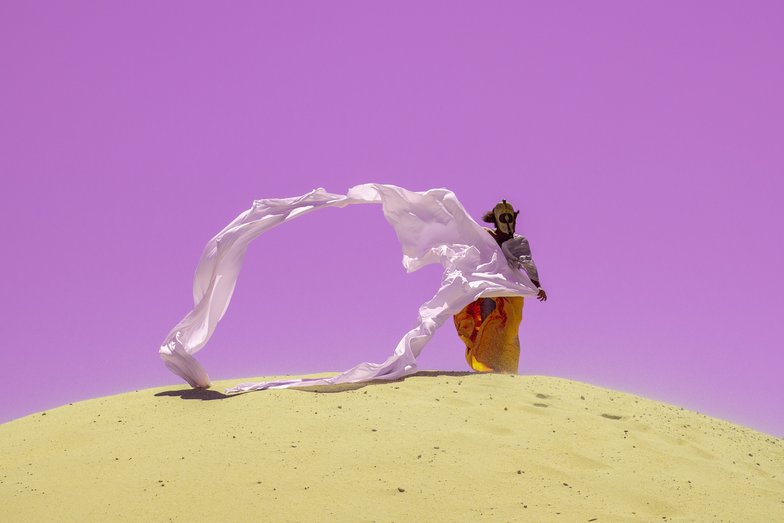 Photograph of a person in the desert. She wears a mask and long robes that are blown into the air by the wind.