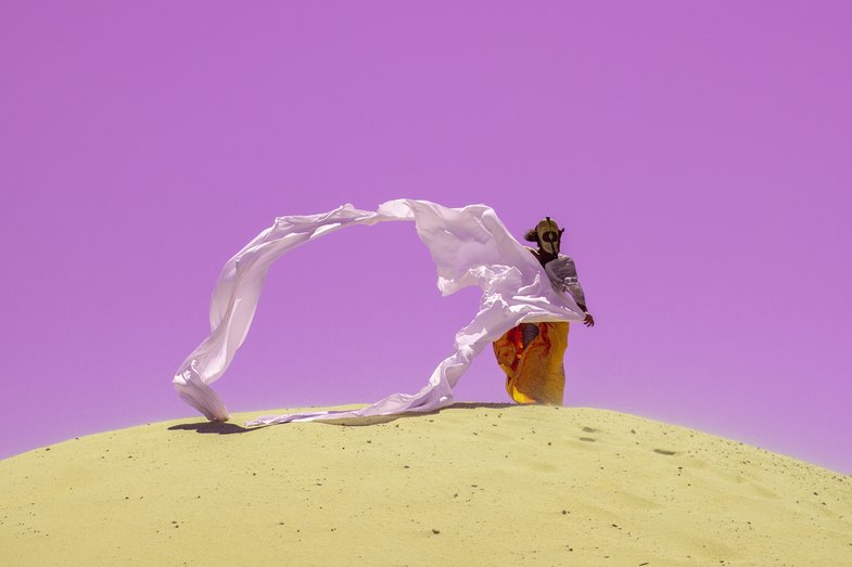 Fotografie einer Person in der Wüste. Sie trägt eine Maske und lange Gewänder, die vom Wind in die Luft getragen werden. 