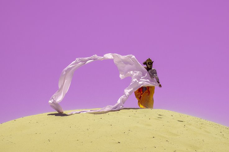 Photograph of a person in the desert. She wears a mask and long robes that are blown into the air by the wind.