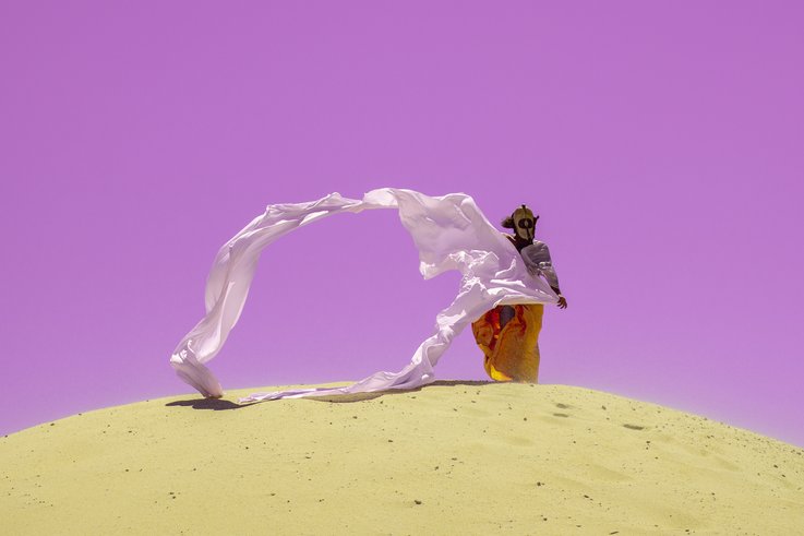 Fotografie einer Person in der Wüste. Sie trägt eine Maske und lange Gewänder, die vom Wind in die Luft getragen werden. 