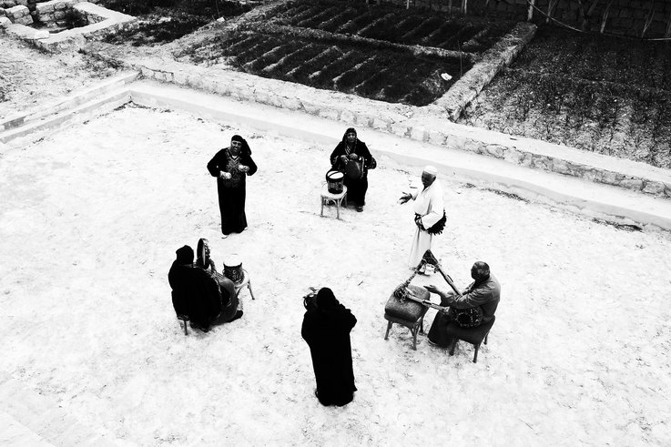 Bird's-eye view photograph of a group of people playing percussion and stringed instruments in a circle. They stand on a sandy square next to cultivated fields framed by small walls.