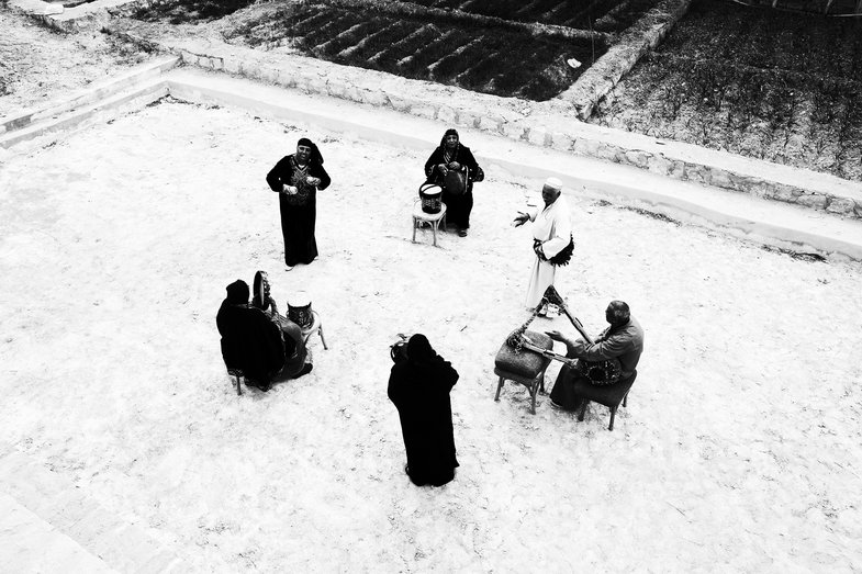 Bird's-eye view photograph of a group of people playing percussion and stringed instruments in a circle. They stand on a sandy square next to cultivated fields framed by small walls.