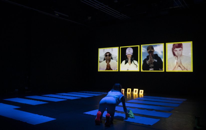 One person rolls up yoga mats in a grid. In the background are images of people from pop culture with iconic folded hands.