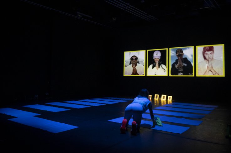 One person rolls up yoga mats in a grid. In the background are images of people from pop culture with iconic folded hands.