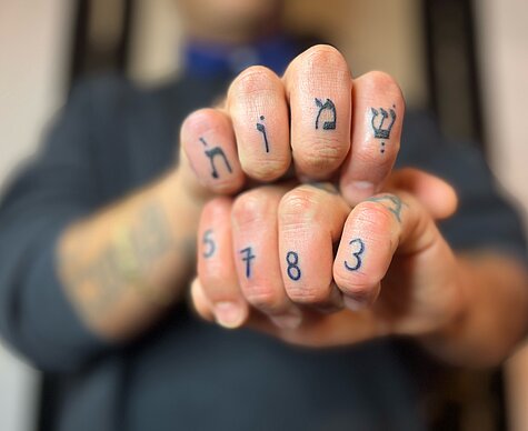 Fotographie zweier gefalteter Hände übereinander. Die Mittelglieder der Finger sind tätowiert. Auf Hebräisch das Wort Namen und die Zahlenreihe 5783 