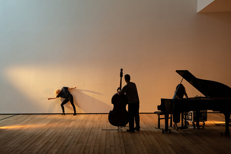 3.	A dancer dances against a wall in the back, with a double bassist and someone playing a grand piano. 