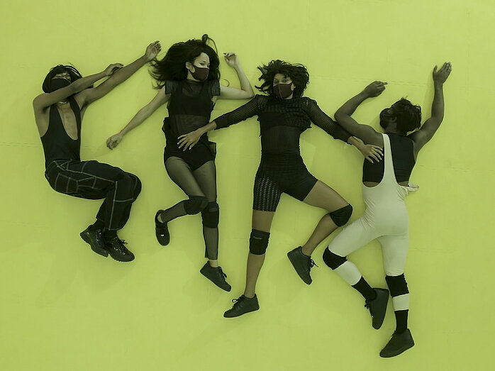 Four people lie on the floor next to each other all wearing black face masks.