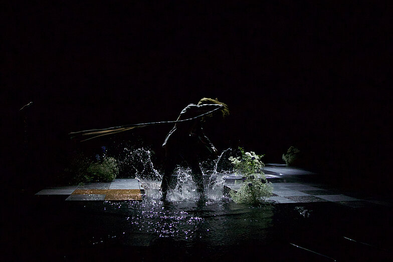 On a stage, a person stands in a puddle. The person twirls around with a long whip.