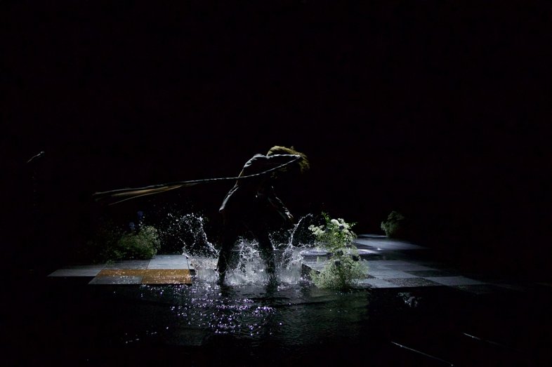 On a stage, a person (Kat Válastur) stands in a puddle. The person twirls around with a long whip.