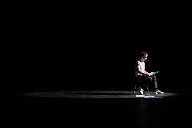 Fotografie von Boris Nikitin auf einer leeren Bühne. Er sitzt auf einem Stuhl und liest einen Text vor.