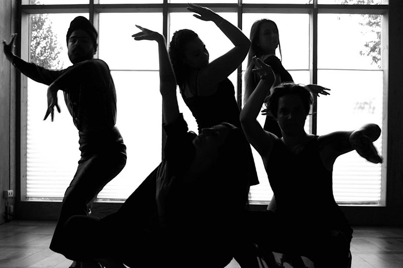 Five dancing people stand in front of a window through which light enters, their faces are barely visible, they are very dark. The photo is a black and white image.