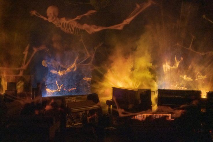 Photograph of a stage on which numerous upright pianos have an open case so that the hammer mechanism is free. Human skeletons hover above them. The foggy room makes the lighting appear dramatic.