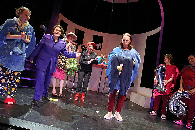 Several people are standing on a stage, their attention is directed towards a person standing somewhat in the foreground, who is holding a pair of trousers in his hand from which a sock is sticking out, reminiscent of an erect penis. Two people on the far right of the picture are holding a silver one and a six as a balloon.