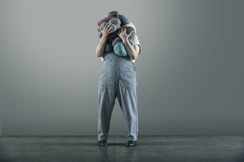 One person is holding so many different coloured round cushions that look a bit like rocks in their arms that their face completely disappears behind them. The person is wearing an overall.