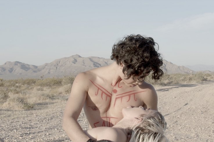 One person holds another person's torso on their lap. The chest is painted with red symbols. The person on the lap licks the other person's chest.