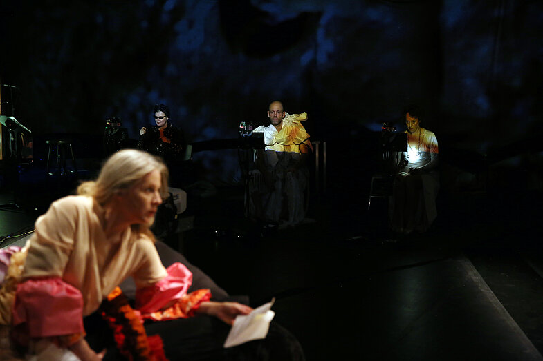 Several performers sit on a stage in the background. In the foreground a woman kneels.