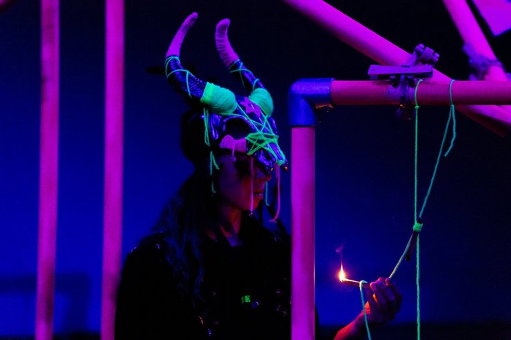 A person sits in the middle of a scaffolding and holds a lit match in his hand. She wears a horned mask.