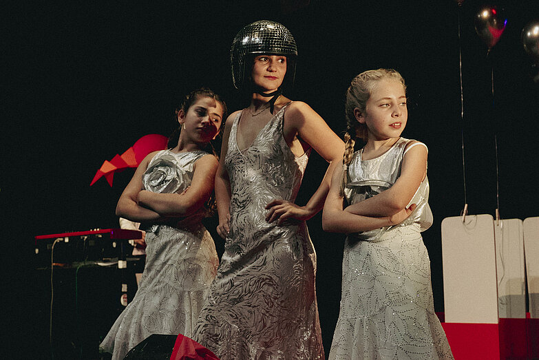 3. 3 People stand in silver costumes. Two younger girls have their arms crossed on either side of a woman who is wearing a silver sparkly helmet in the middle with her arms on her hips.