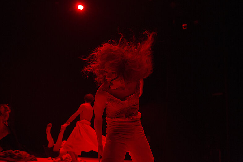 Three characters on an empty stage. In the foreground a person shakes his head, in the background a person is holding another's ankle and hand and turning with her.