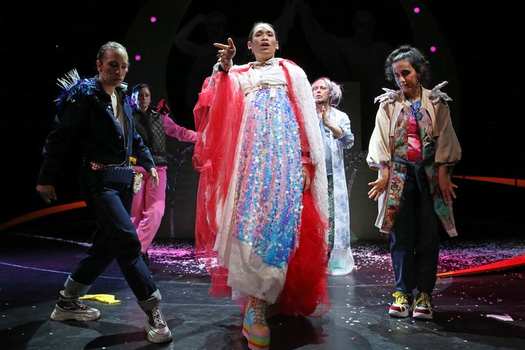 Five people on stage, three in the foreground, two in the second row. The person in the middle walks towards the camera wearing a long, glittery dress. There are many confettis on the stage.