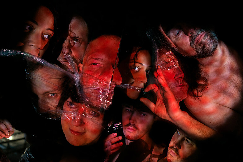 10 mit Frischhaltefolie bedeckte Gesichter sind in einem roten Licht auf ungewöhnliche Weise angeordnet. Auf dem Bild sind drei Hände zu sehen.